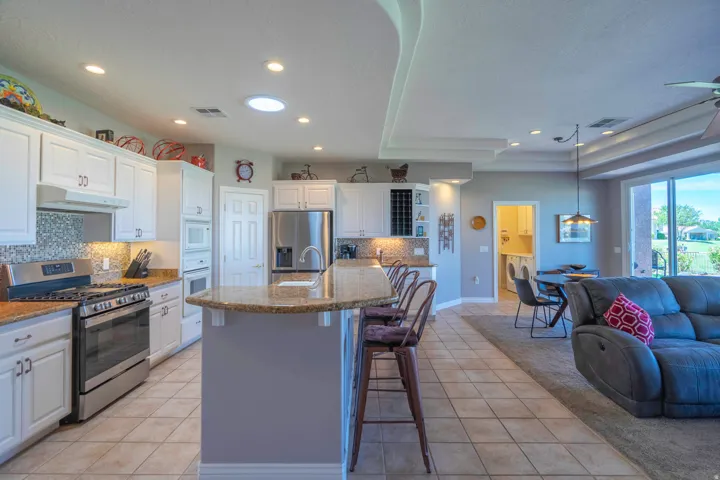 Open-concept kitchen and living area featuring white cabinetry with stainless steel appliances, a large center island with seating, tile flooring throughout, recessed lighting, and expansive windows providing natural light