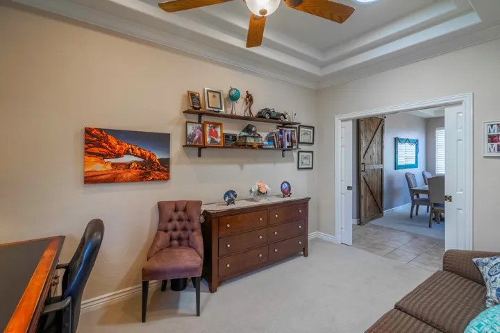 Versatile secondary bedroom featuring coffered ceiling detail, ceiling fan with integrated lighting, built-in shelving with display cubbies, wood-finish dresser, and neutral wall-to-wall carpeting