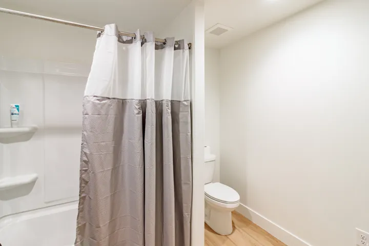 White bathtub with integrated shelving, a chrome shower rod, a white toilet, and wood-finish flooring