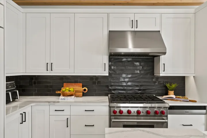 White shaker-style cabinetry, stainless steel range hood, dark textured backsplash, professional-grade gas range, and light-toned countertops