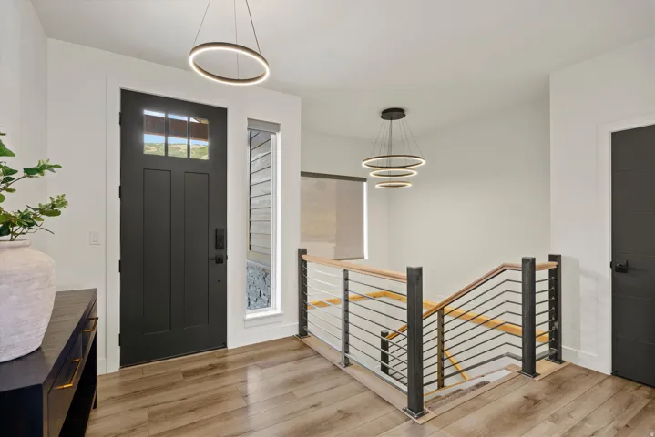 Wood-finish flooring, dark-toned entry door with glass panels, modern circular pendant lighting, wood and metal railing, and white wall finishes