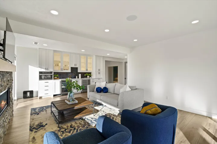 Integrated living space featuring a stone-clad fireplace, light wood-finish flooring, recessed lighting, white built-in cabinetry with glass-front uppers, and stainless steel beverage refrigerators