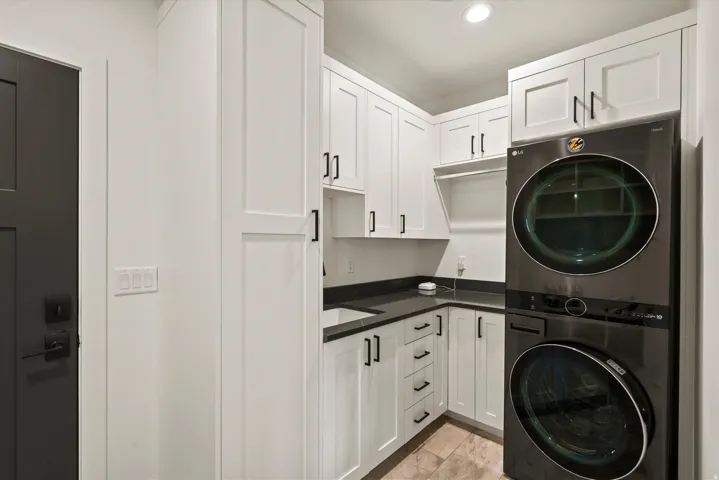 Laundry area with white shaker-style cabinetry, dark countertops, an undermount sink, and a stacked washer and dryer