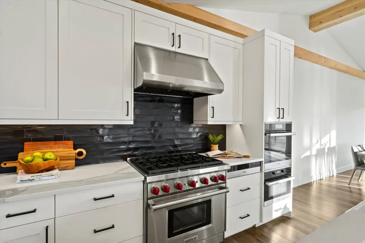 Stainless steel range with matching hood, white shaker cabinetry, dark subway tile backsplash, light wood-finish flooring, and exposed ceiling beams