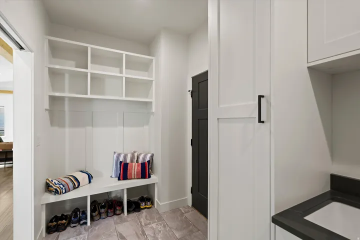 Built-in storage bench with open shelving, dark-finish interior door, light-toned tile flooring, white shaker-style cabinetry, and dark countertop with an integrated sink
