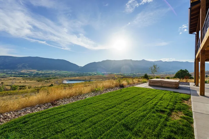 Expansive mountain backdrop, manicured lawn, concrete patio, and upper-level balcony with metal railing