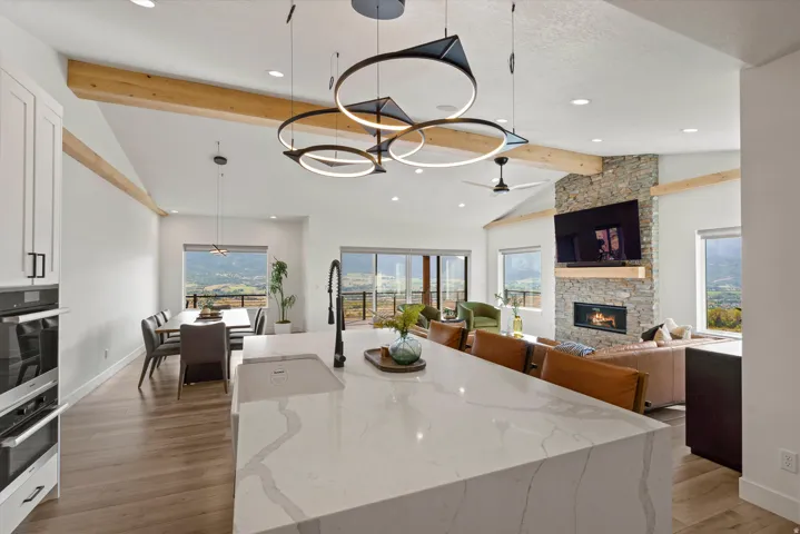 Vaulted ceiling with exposed wood beams, stone fireplace, large sliding glass doors, white shaker cabinetry, and a large kitchen island with integrated sink