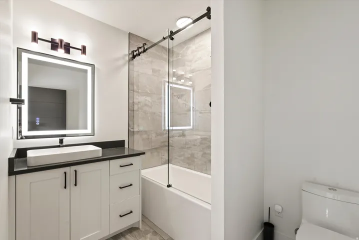 White shaker-style vanity with dark countertop, vessel sink, and an illuminated mirror