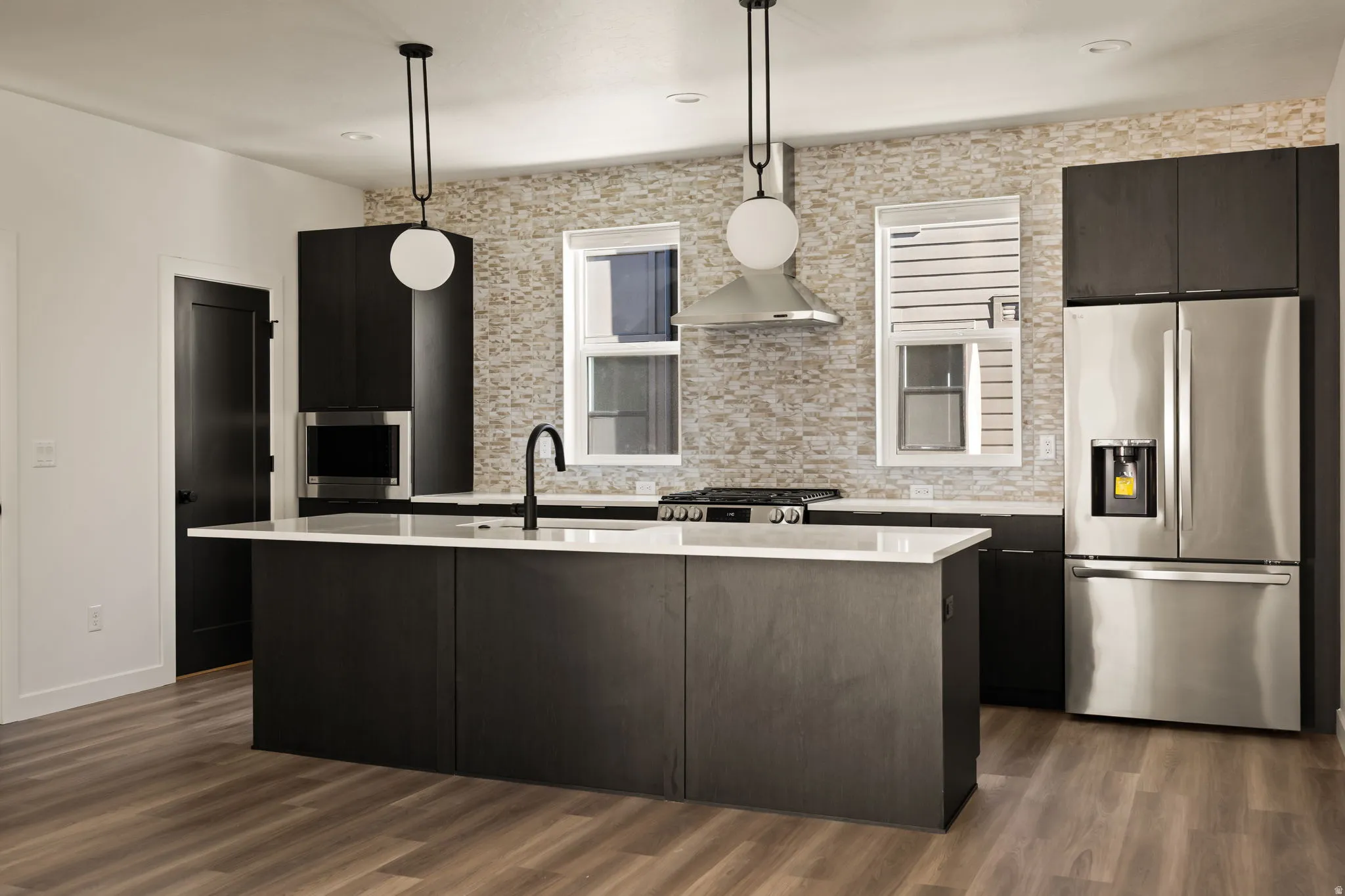 Modern kitchen featuring an expansive center island, integrated stainless steel appliances, dark wood-finish cabinetry, a textured tile backsplash, and wood-finish flooring