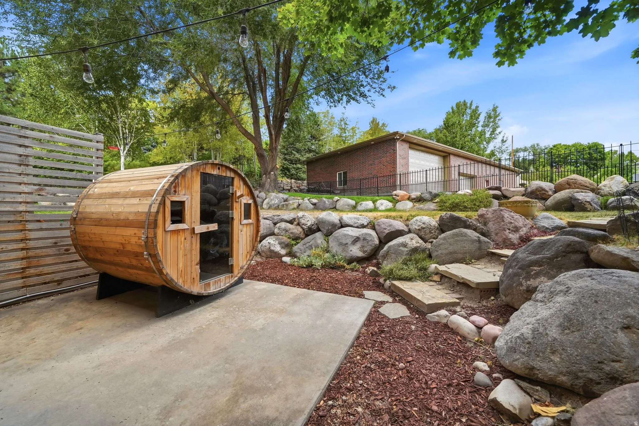 Barrel sauna with clear door, concrete patio, slatted privacy wall, and rock landscaping