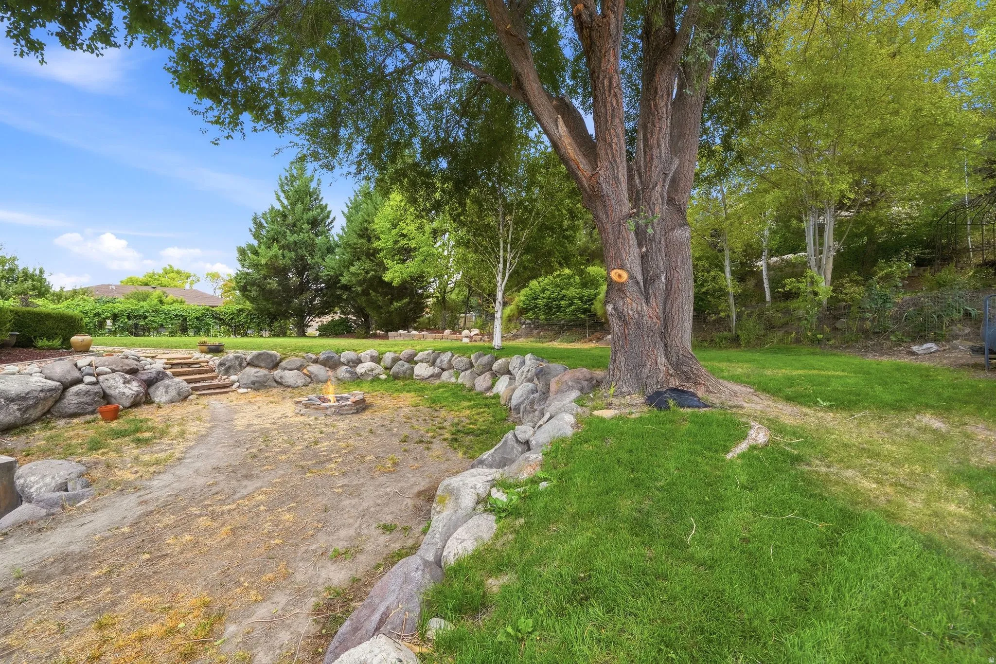 Landscaped backyard featuring a stone retaining wall, mature trees, a built-in fire pit, and stone steps