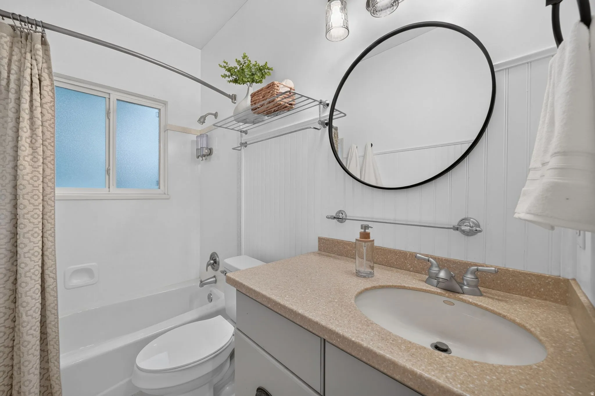 Bathroom featuring a built-in bathtub with shower, a vanity with an integrated sink and light-colored countertop, a wall-mounted round mirror, and beadboard wall paneling