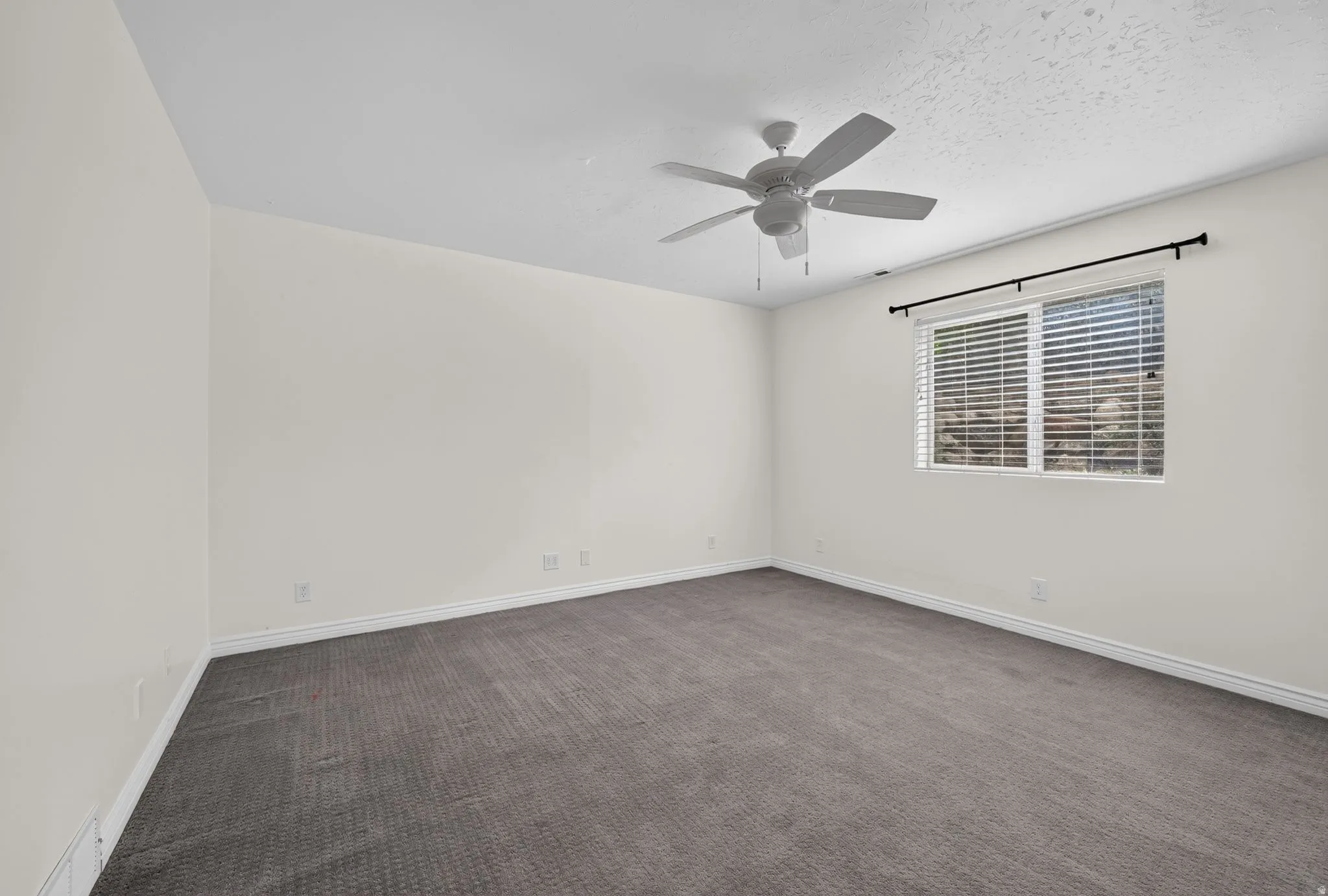 Spacious room featuring neutral carpeting, white walls, a ceiling fan, and a window with horizontal blinds and a black curtain rod
