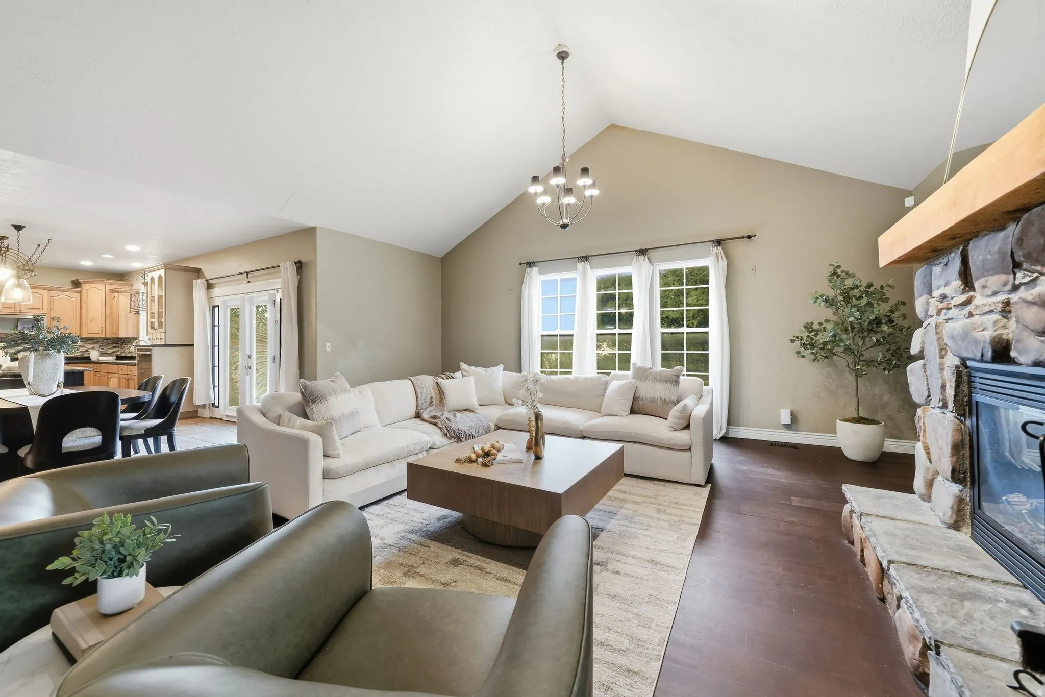 Vaulted ceiling featuring a contemporary chandelier, stone fireplace with a substantial wood mantel, wood-finish flooring, and multiple windows