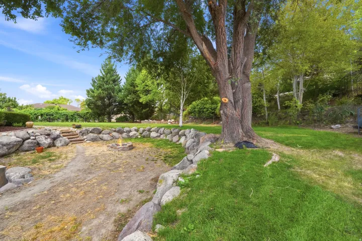 Landscaped backyard featuring a stone retaining wall, mature trees, a built-in fire pit, and stone steps
