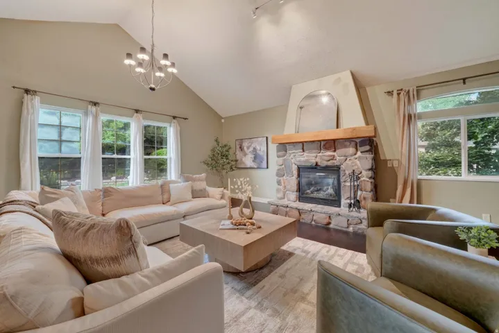 Vaulted ceiling with an attached chandelier, stone-surround fireplace with a wood mantel, and multiple windows