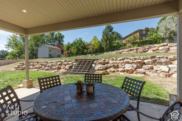 Covered patio with a round slate patio table, overlooking a tiered stone retaining wall and stairs leading to upper lawn and newer storage shed.