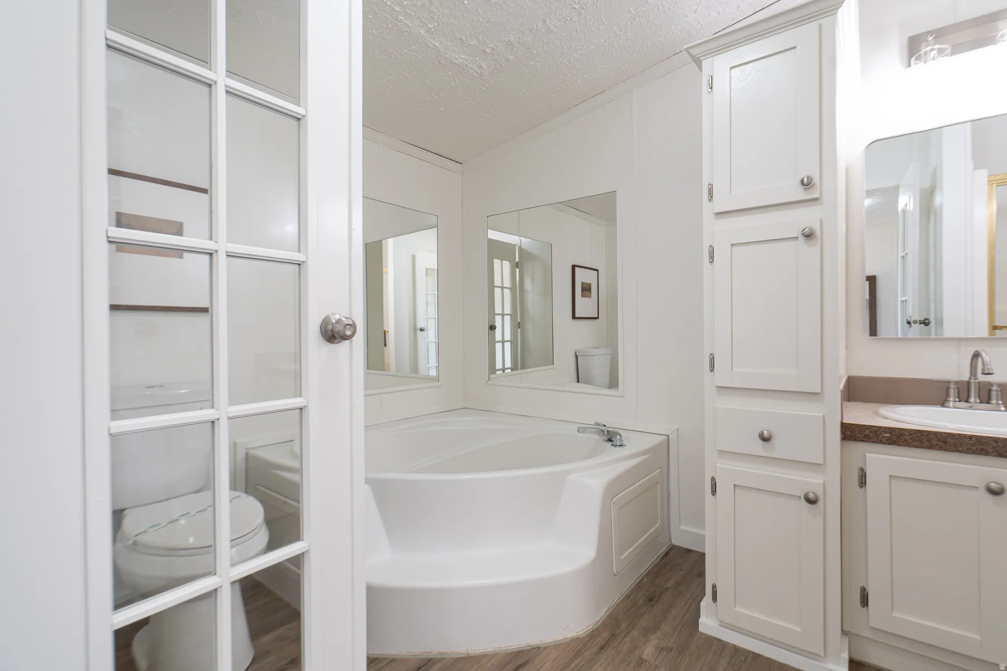 Enclosed water closet, corner soaking tub, wood-finish flooring, built-in linen cabinet, and a vanity with an undermount sink and stone-finish countertop
