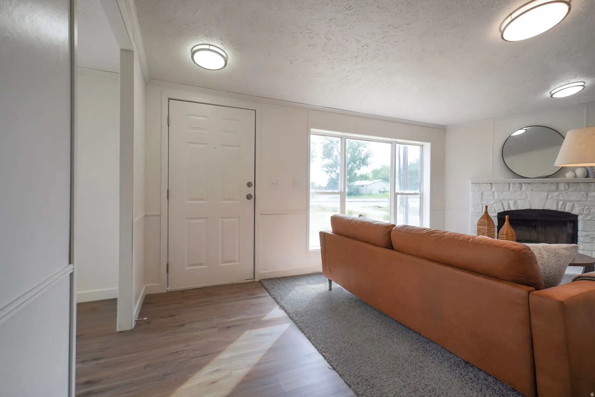 Wood-finish flooring, white paneled door, fireplace with stone surround, flush-mount ceiling lighting, and large windows