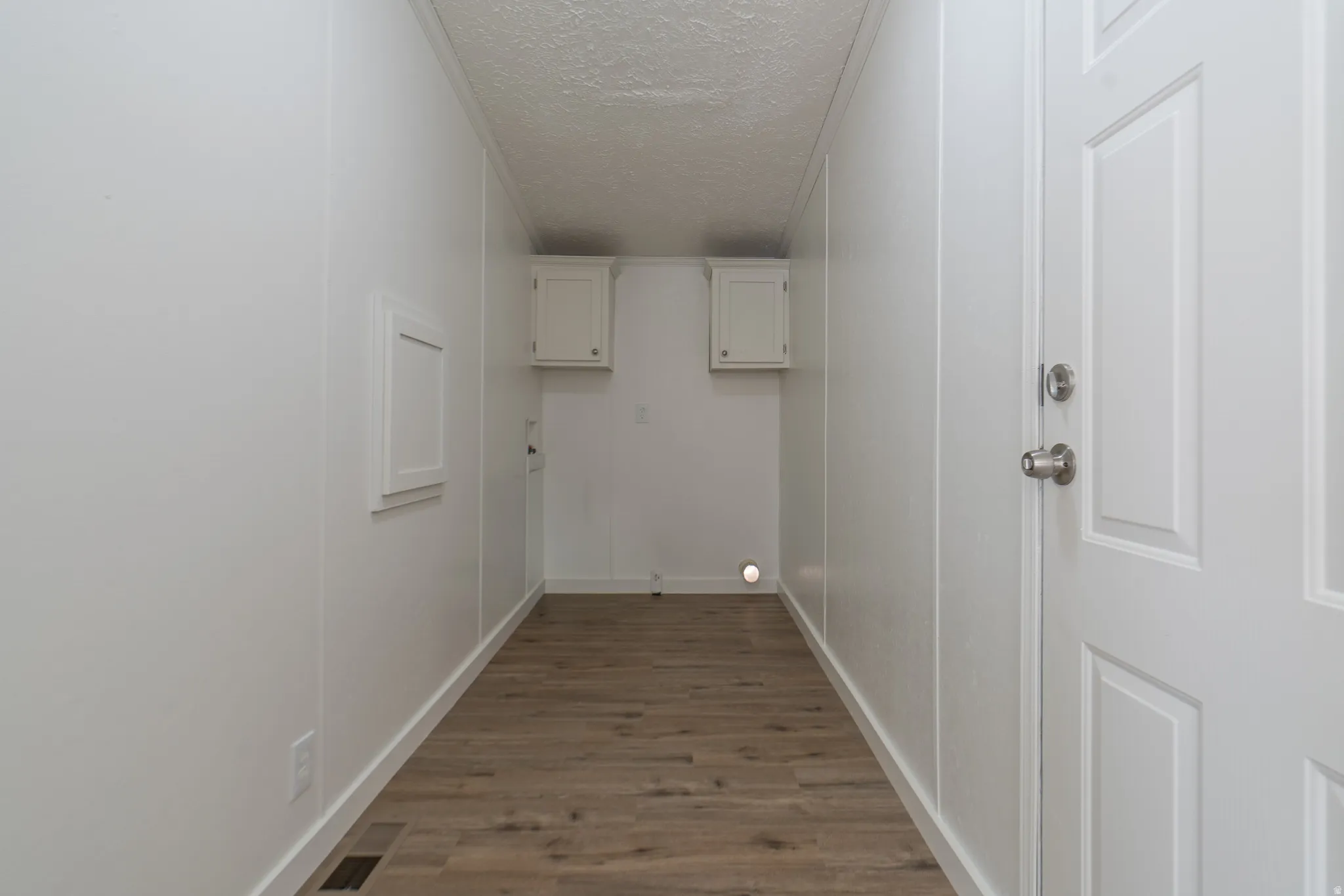 Wood-finish flooring with white wall paneling, two white upper cabinets, and a white paneled door with brushed nickel hardware