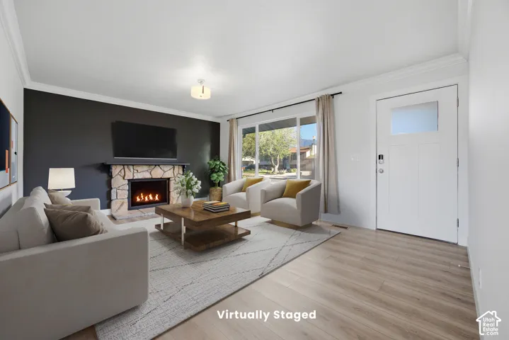 Wood-finish flooring, stone-clad fireplace, large front-facing window, white crown molding, and a modern entry door with frosted glass insert