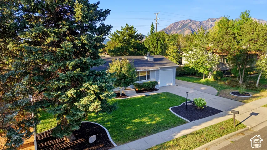 Single-story residence with an attached garage, featuring a paved driveway, mature trees, manicured lawn, and mountain scenery