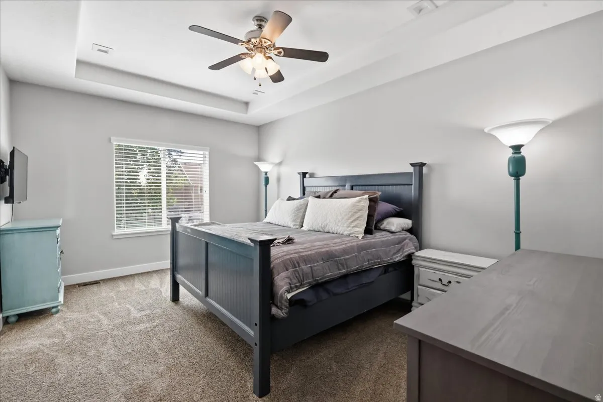 Tray ceiling with recessed lighting, ceiling fan with integrated lighting, carpet flooring, and a large window with blinds