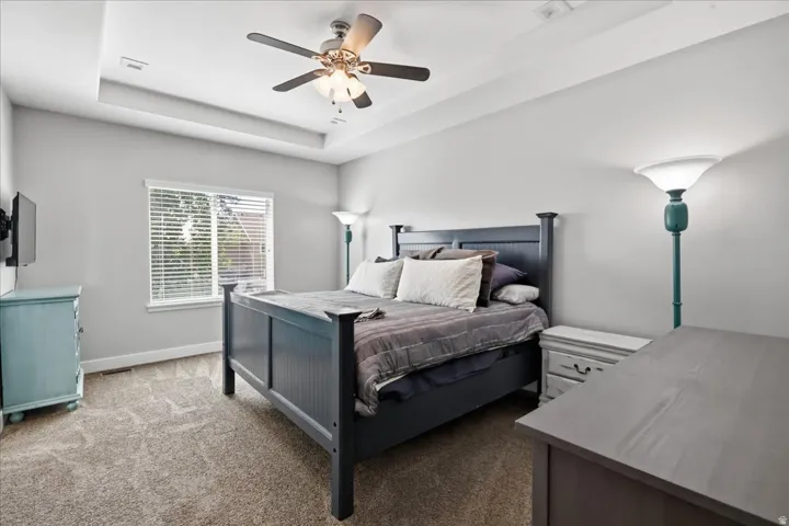 Tray ceiling with recessed lighting, ceiling fan with integrated lighting, carpet flooring, and a large window with blinds