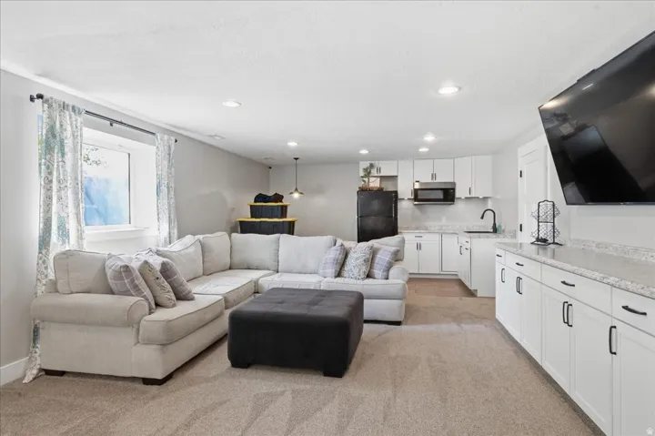 Finished space featuring carpet flooring, recessed lighting, a built-in wet bar with white cabinetry and light-toned countertops, and a kitchen area with white cabinetry, a black sink, and black appliances