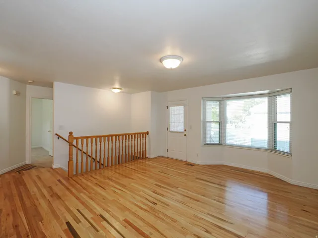 Hardwood flooring throughout, bay window with blinds, white walls, wood baluster railing, and ceiling-mounted light fixtures