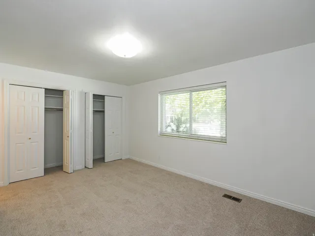 Neutral-toned carpeting, dual bi-fold door closets, flush-mount ceiling lighting, and a large window with blinds