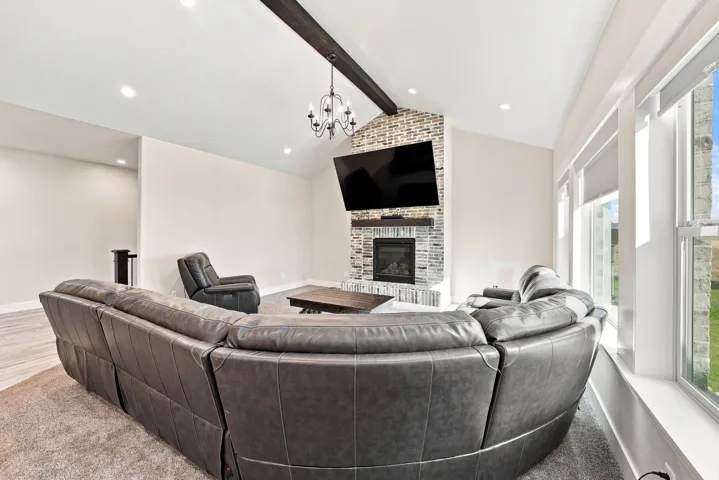 Vaulted ceiling with exposed wood beam, stone fireplace with a built-in electric insert, large windows with roller shades, recessed lighting, and wood-finish flooring