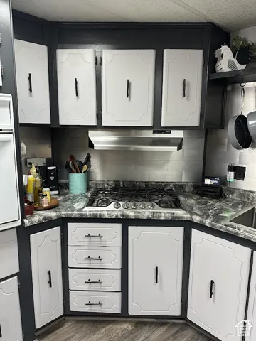 Gray and white cabinetry with dark hardware, gas range with stainless steel hood, dark marble-look countertops, and wood-finish flooring