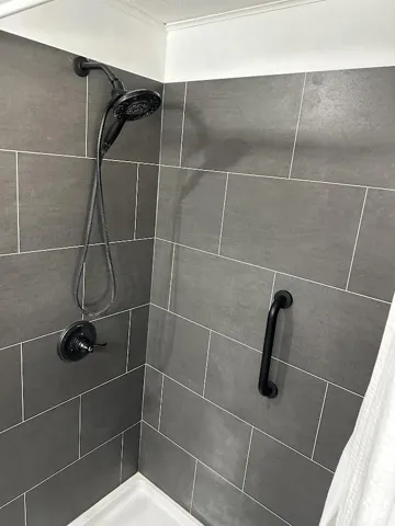 Shower with dark grey tile surround, white grout, black shower fixtures, and a black grab bar