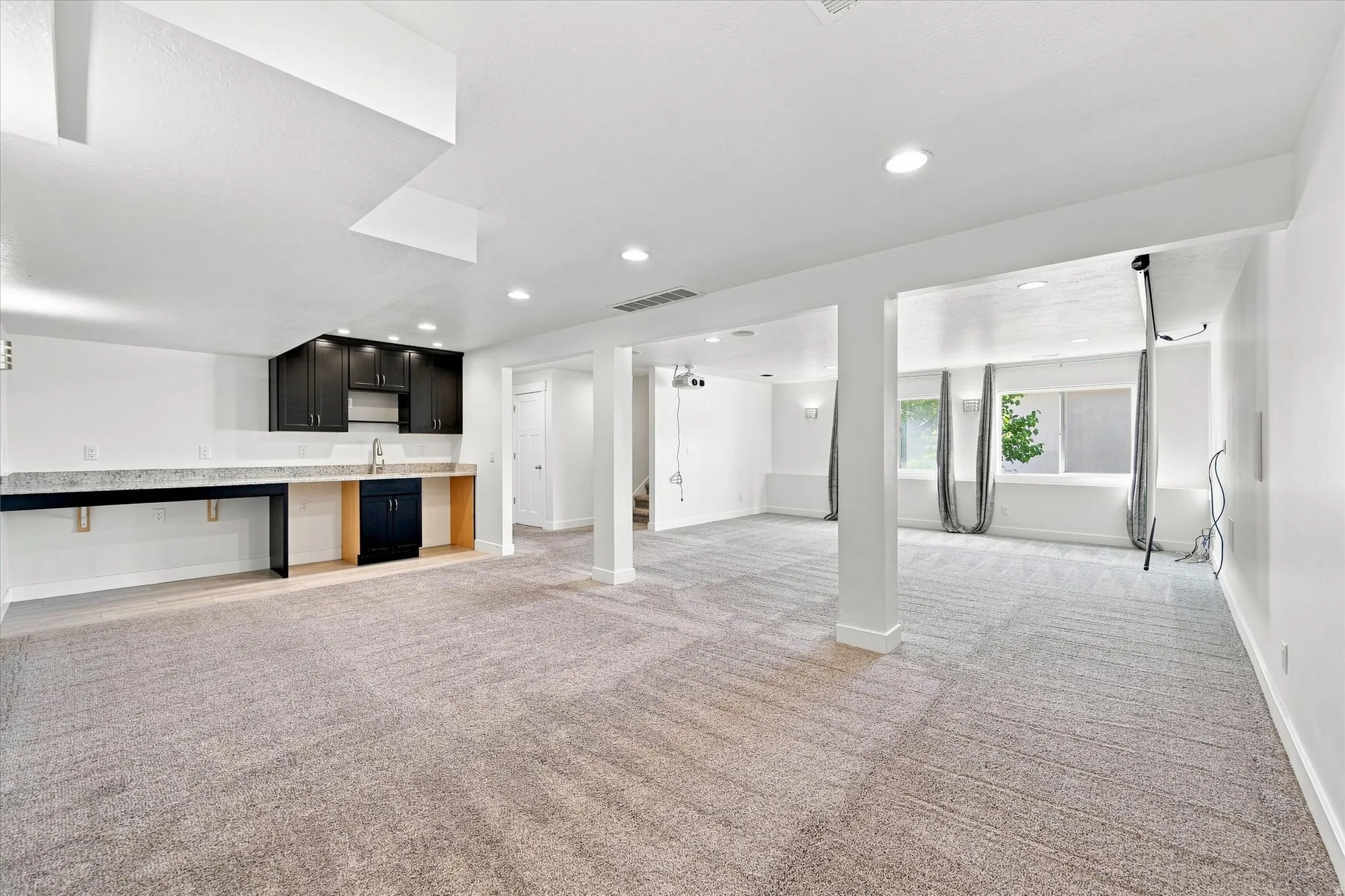 Spacious finished room with recessed lighting, light-colored carpeting, dark cabinetry with light-toned countertops, and multiple windows