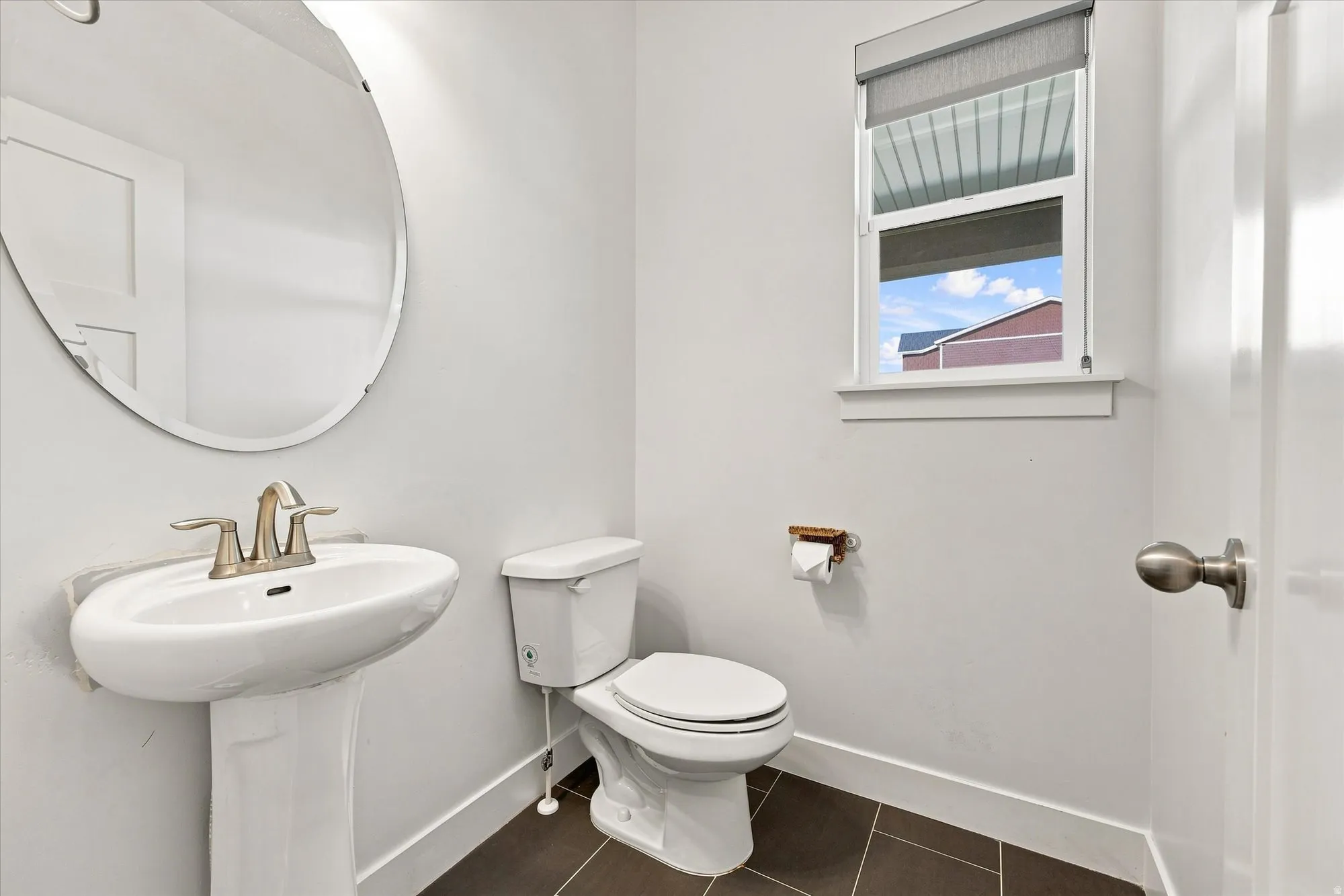 Pedestal sink with brushed nickel faucet, oval wall mirror, white toilet, and dark tile flooring