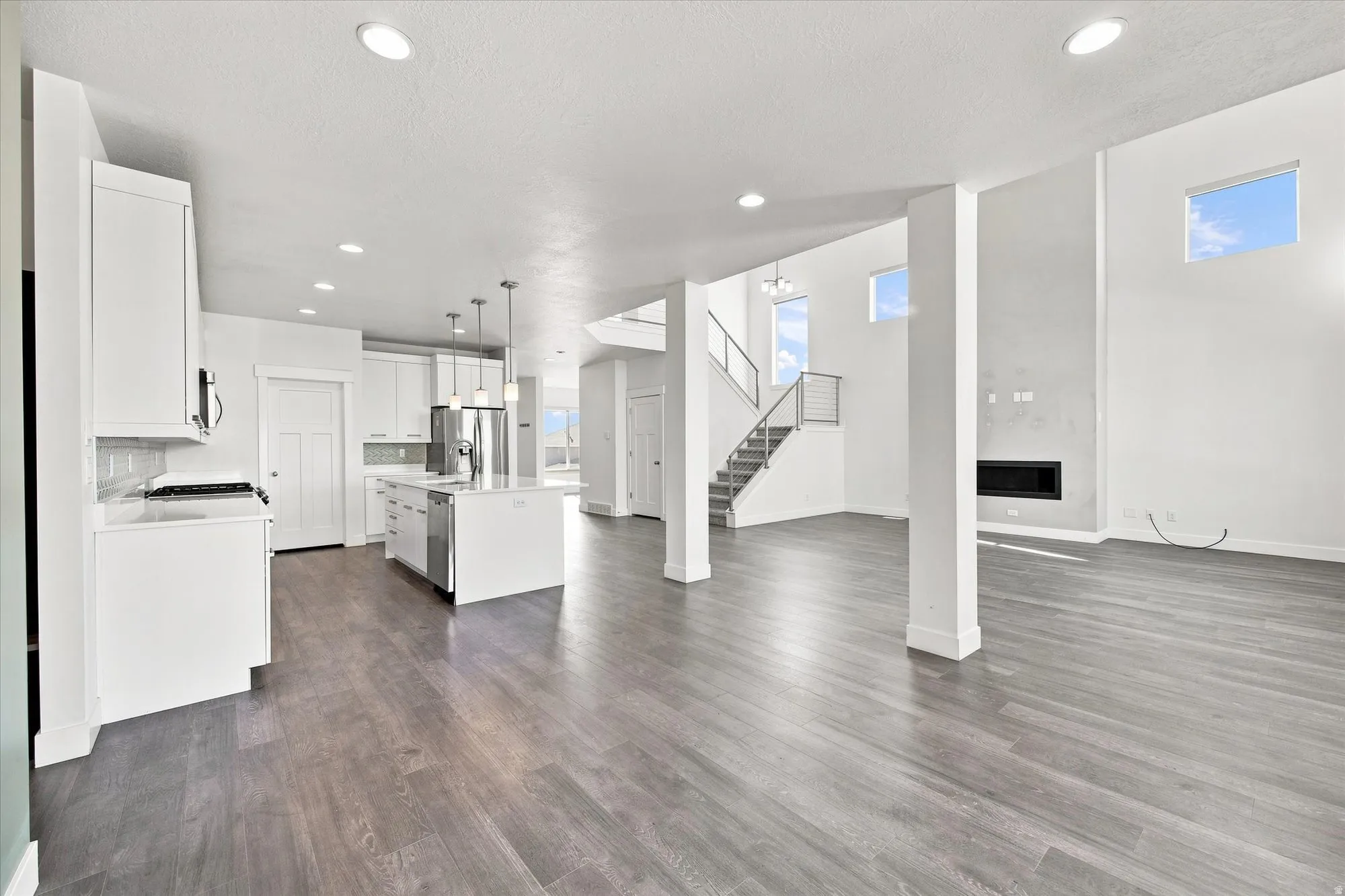 Open-concept living space featuring wood-finish flooring, a modern kitchen with a central island, a built-in fireplace, and a staircase with a cable railing