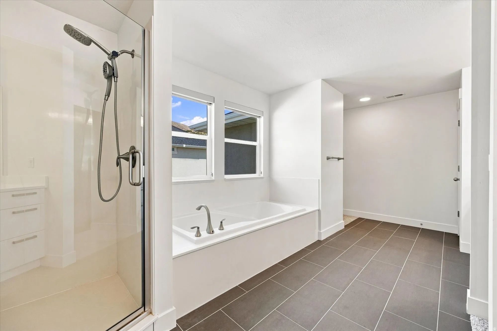 Expansive bathroom featuring a walk-in shower with glass enclosure, a large soaking tub with dual windows, and dark gray rectangular floor tiles