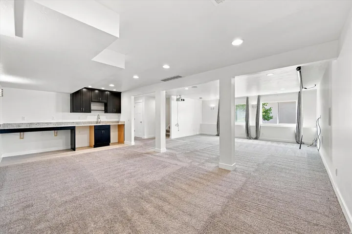 Spacious finished room with recessed lighting, light-colored carpeting, dark cabinetry with light-toned countertops, and multiple windows