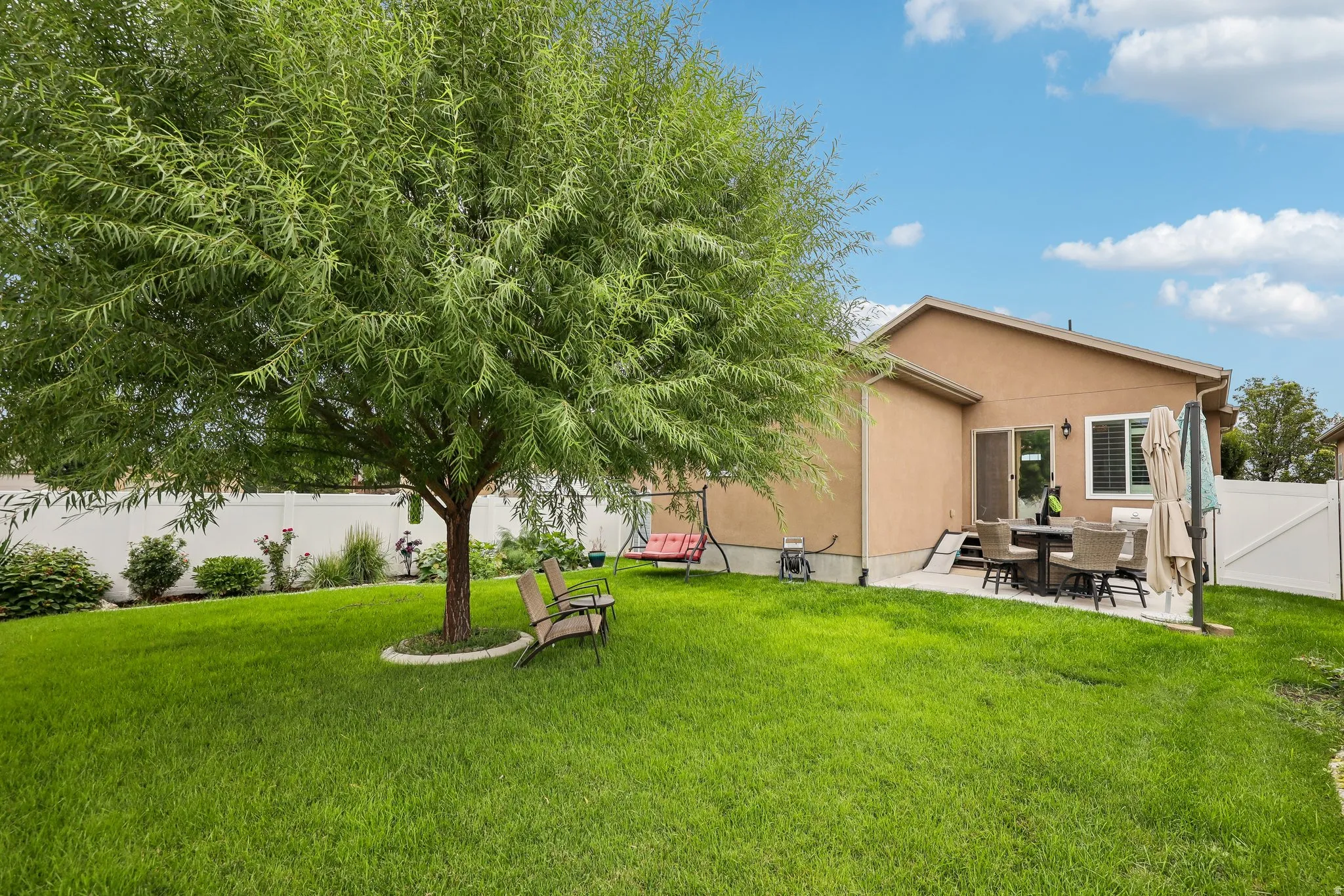 Expansive green lawn, mature tree, concrete patio, and stucco exterior with white fencing