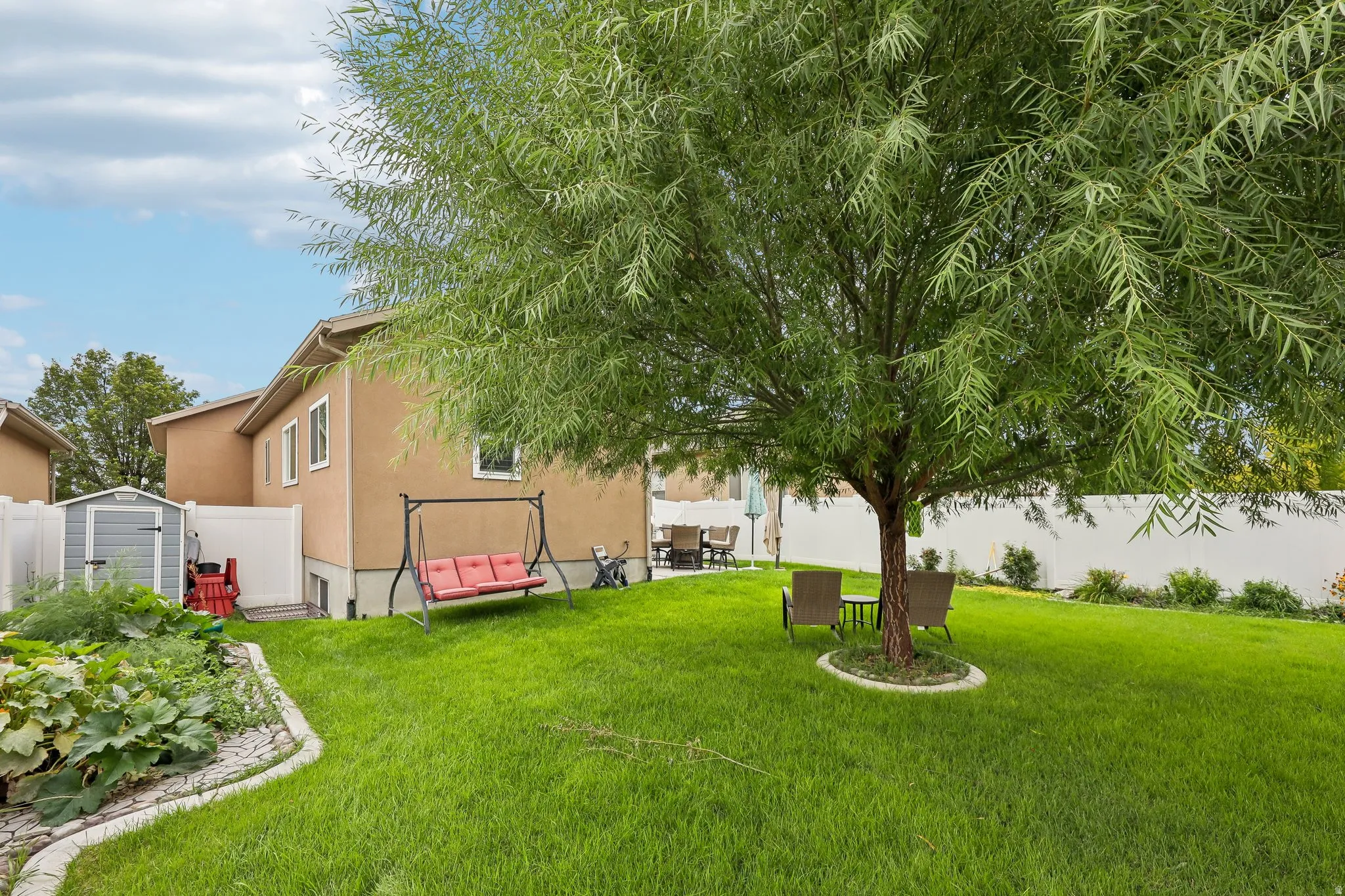 Expansive backyard featuring a mature tree, a manicured lawn, a garden bed with stone edging, a stucco exterior, and a privacy fence