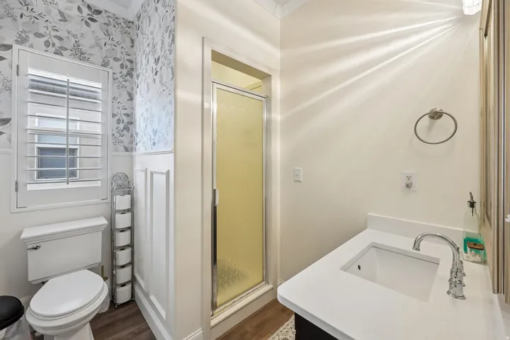 Vanity with integrated sink and chrome faucet, glass-enclosed shower, white wainscoting, and wood-finish flooring