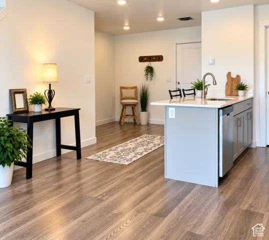 Wood-finish flooring throughout, recessed lighting, kitchen island with integrated sink, stainless steel dishwasher, and light gray cabinetry
