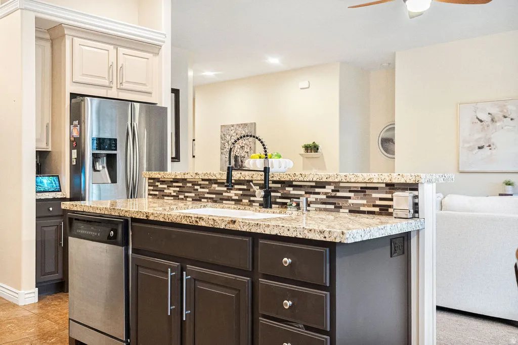 Granite-style countertop, stainless steel appliances, dark wood-finish cabinetry, tile backsplash, and a ceiling fan with integrated lighting