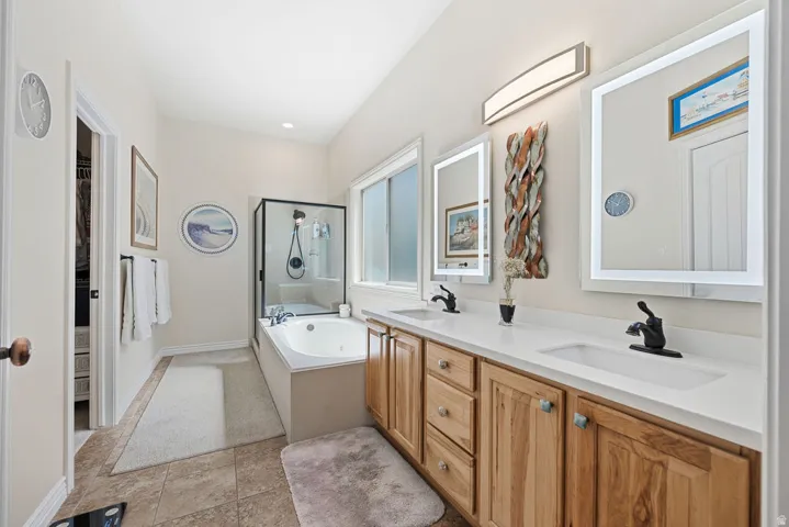 Dual vanity with integrated sinks, light wood cabinetry, illuminated mirrors, built-in bathtub, and a glass-enclosed shower