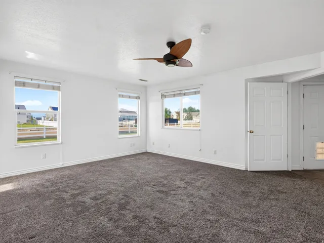 White interior walls, dark gray carpet, two windows with white trim, and a ceiling fan with wood-finish blades