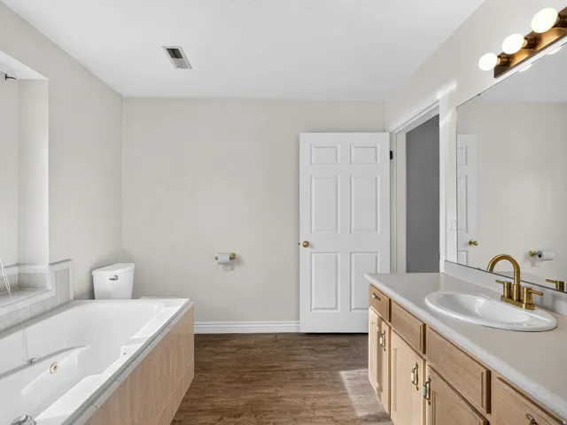 Soaking tub, wood-finish flooring, vanity with integrated sink, brass-finish faucet, and a large wall mirror