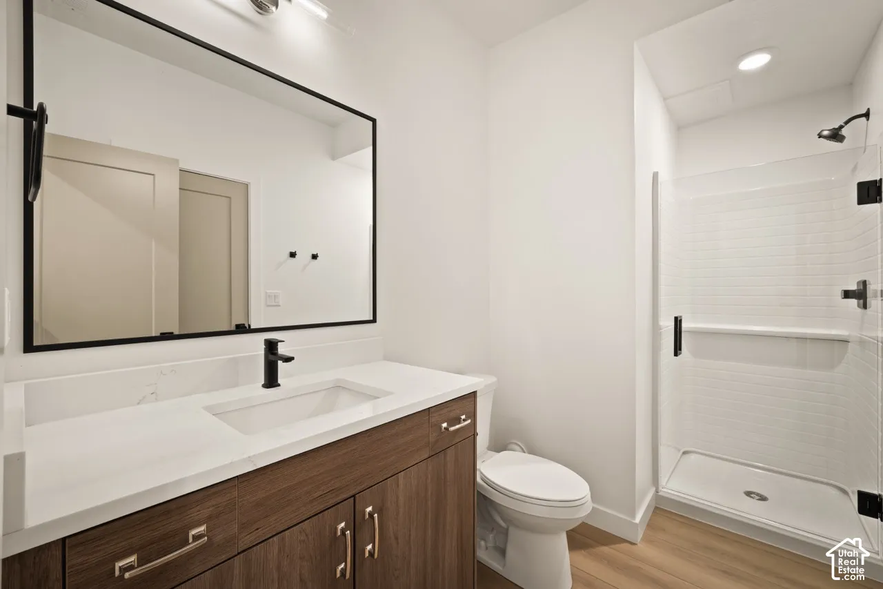 Wood-finish flooring, white vanity countertop, wood cabinetry, modern black fixtures, and a glass-enclosed shower with white tile