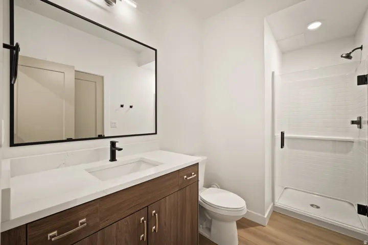 Wood-finish flooring, white vanity countertop, wood cabinetry, modern black fixtures, and a glass-enclosed shower with white tile