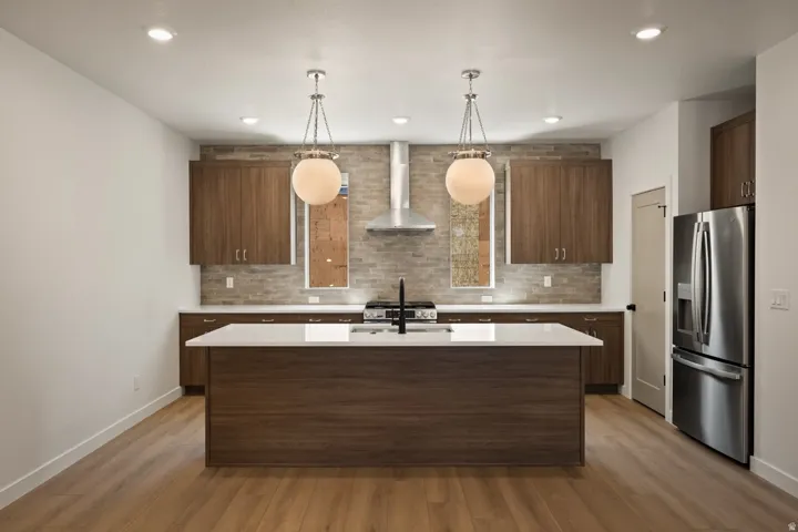 Kitchen featuring a central island with white countertop, wood-finish cabinetry, stainless steel appliances, brick-patterned backsplash, and pendant lighting